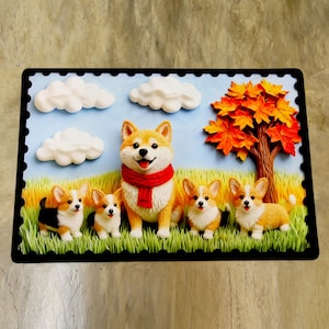 May include: A rectangular decorative piece depicting a corgi family. The central corgi wears a red scarf, surrounded by smaller corgis in a grassy field. White clouds float in a blue sky, with an autumn tree in the background.