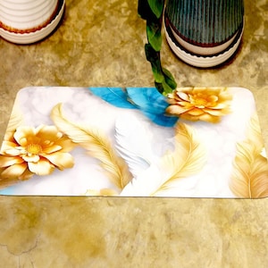 May include: Rectangular floor mat with a floral and feather design. The mat has a white background with yellow flowers, gold feathers, and blue accents. The mat is likely made of a durable, washable material.