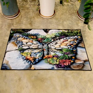 May include: Rectangular doormat with a butterfly design. The butterfly wings are made of stones, succulents, and greenery, with a black border. The design incorporates various colours, including green, brown, orange, and purple.