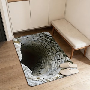 May include: A rectangular floor mat featuring a detailed image of a stone well. The well's interior is dark, contrasting with the lighter stone bricks. Two pairs of beige slippers are placed on the wooden floor next to the mat.