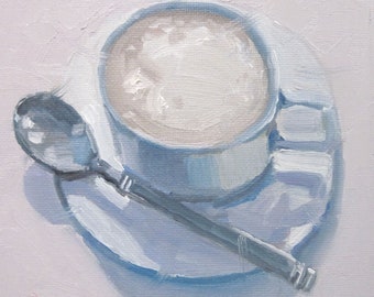 Könnte beinhalten: Eine weiße Kaffeetasse aus Keramik mit einer hellbraunen Flüssigkeit und weißem Schaum darin, die auf einem weißen Unterteller mit einem silbernen Löffel liegt.