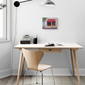 K&ouml;nnte beinhalten: Ein quadratisches Gem&auml;lde eines Ladens mit roter Markise h&auml;ngt an einer wei&szlig;en Wand. Vor der Wand stehen ein Holztisch mit Schublade und ein Holzstuhl mit Metallbeinen. &Uuml;ber dem Schreibtisch ist eine schwarze Lampe an der Wand befestigt.
