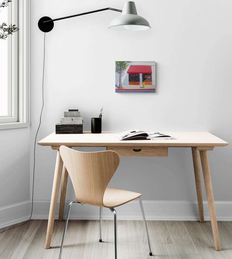 K&ouml;nnte beinhalten: Ein quadratisches Gem&auml;lde eines Ladens mit roter Markise h&auml;ngt an einer wei&szlig;en Wand. Vor der Wand stehen ein Holztisch mit Schublade und ein Holzstuhl mit Metallbeinen. &Uuml;ber dem Schreibtisch ist eine schwarze Lampe an der Wand befestigt.
