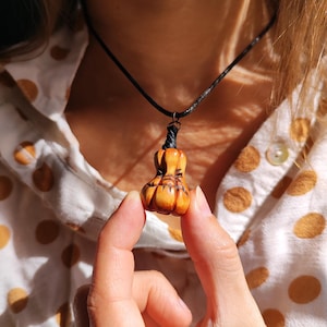 May include: A small, hand-carved wooden pendant shaped like a pumpkin with a smooth, polished orange-brown finish and dark etched details. The pendant hangs from a simple black cord necklace, featuring a dark stem and segmented body.