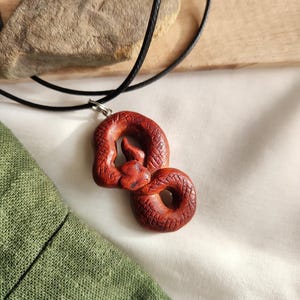 May include: A handcrafted red snake pendant on a black cord necklace. The snake is coiled with detailed scales and a small head. The pendant is attached to a black cord necklace, suitable for everyday wear.