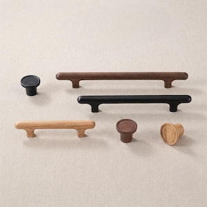 May include: A collection of cabinet hardware including two long horizontal bar pulls in dark and black wood, one shorter light wood bar pull, and three round knobs in black, dark wood, and light wood finishes with smooth, cylindrical shapes.
