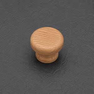 May include: A round cabinet knob crafted from light brown wood with a visible natural grain pattern. The knob features a flat circular top surface that tapers down to a smaller cylindrical base, showcasing a smooth, polished finish throughout.