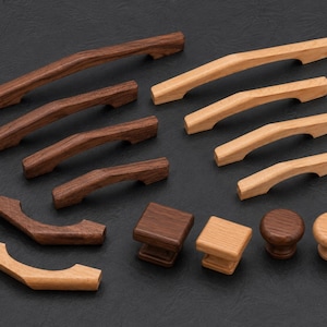 May include: A collection of wooden cabinet hardware including various sizes of arched drawer pulls, square knobs, and round knobs. The hardware is crafted from two distinct wood tones, featuring a dark brown walnut finish and a light natural oak finish.