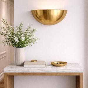 May include: A gold-colored, half-moon shaped wall sconce illuminates a white wall. Below, a marble-topped table with a wooden frame holds a vase of white flowers, books, and a small gold bowl. The scene evokes a sense of modern elegance.