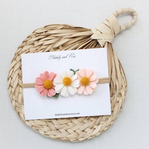 May include: A white card with the text "Blakeby and Rae" and the website "blakeleyandrae.etsy.com" is displayed on a woven straw mat. A felt flower headband with three flowers is on the card. The flowers are pink, white, and yellow.