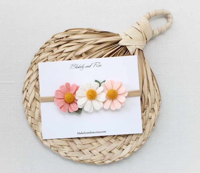 May include: A white card with the text "Blakeby and Rae" and the website "blakeleyandrae.etsy.com" is displayed on a woven straw mat. A felt flower headband with three flowers is on the card. The flowers are pink, white, and yellow.
