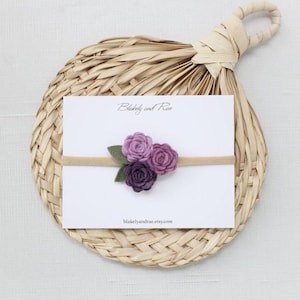 May include: A purple felt flower headband with three flowers in shades of purple and green leaves. The headband is on a white card with the text "Blakeley and Rae" and "blakeleyandrae.etsy.com".