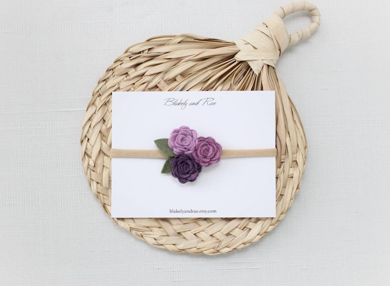 May include: A purple felt flower headband with three flowers in shades of purple and green leaves. The headband is on a white card with the text "Blakeley and Rae" and "blakeleyandrae.etsy.com".