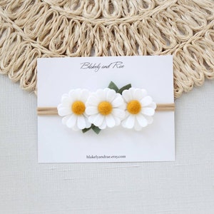 May include: A white felt headband with three white felt daisies with yellow centers and green felt leaves. The headband is on a white card with the text "Blakely and Rae" and "blakelyandrae.etsy.com".
