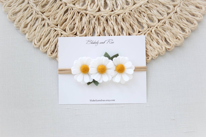 May include: A white felt headband with three white felt daisies with yellow centers and green felt leaves. The headband is on a white card with the text "Blakely and Rae" and "blakelyandrae.etsy.com".
