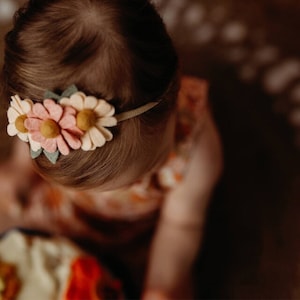 May include: A baby girl wearing a headband with felt flowers in shades of pink, white, and yellow. The headband is made of a cream-colored fabric.