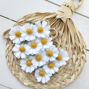 May include: A set of felt daisy hair clips in white with yellow centers and green leaves. The clips are arranged on a woven straw fan.
