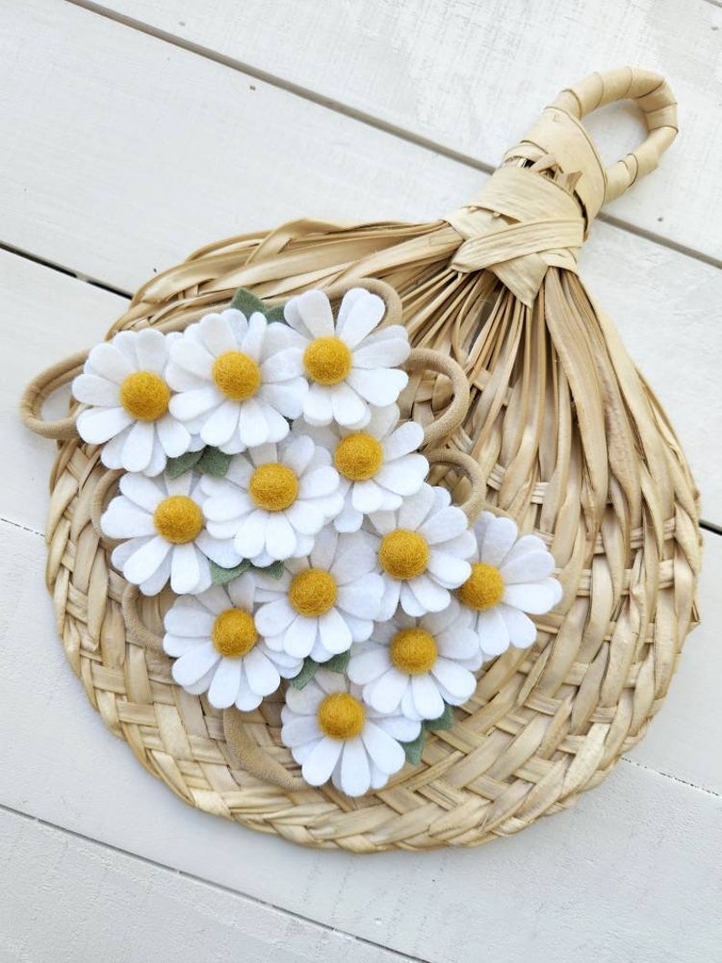 May include: A set of felt daisy hair clips in white with yellow centers and green leaves. The clips are arranged on a woven straw fan.