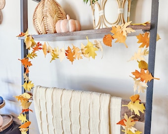 Pode incluir: Uma guirlanda decorativa de folhas de outono em tons de amarelo, laranja e creme, pendurada em uma prateleira de escada cinza escuro. A guirlanda é feita de folhas de feltro. Uma manta creme está pendurada na prateleira da escada. Uma cesta de vime e uma abóbora rosa estão na prateleira.