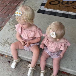 May include: Two toddlers wearing pink off-the-shoulder tops and floral print bottoms. Both girls are wearing flower headbands. The girl on the left is holding a small brown leaf.