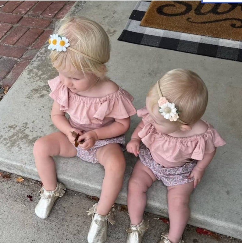 May include: Two toddlers wearing pink off-the-shoulder tops and floral print bottoms. Both girls are wearing flower headbands. The girl on the left is holding a small brown leaf.
