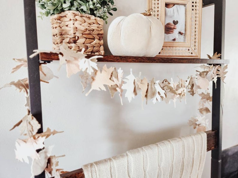 May include: A white pumpkin, a potted green plant, and a framed photo on a wooden shelf with a brown felt leaf garland hanging below the shelf.