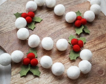 May include: A festive garland featuring white felt balls interspersed with clusters of red berries and green holly leaves. The garland is arranged in a circular pattern on a wooden surface, creating a seasonal decorative element.