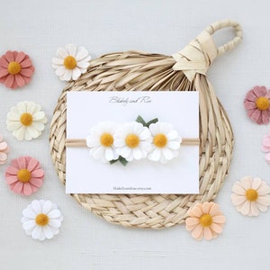 May include: A white felt headband with three white felt daisies and green felt leaves. The headband is on a white card with the text "Blakeley and Rae" and "blakeleyandrae.etsy.com". There are several felt daisies in various colors around the card.
