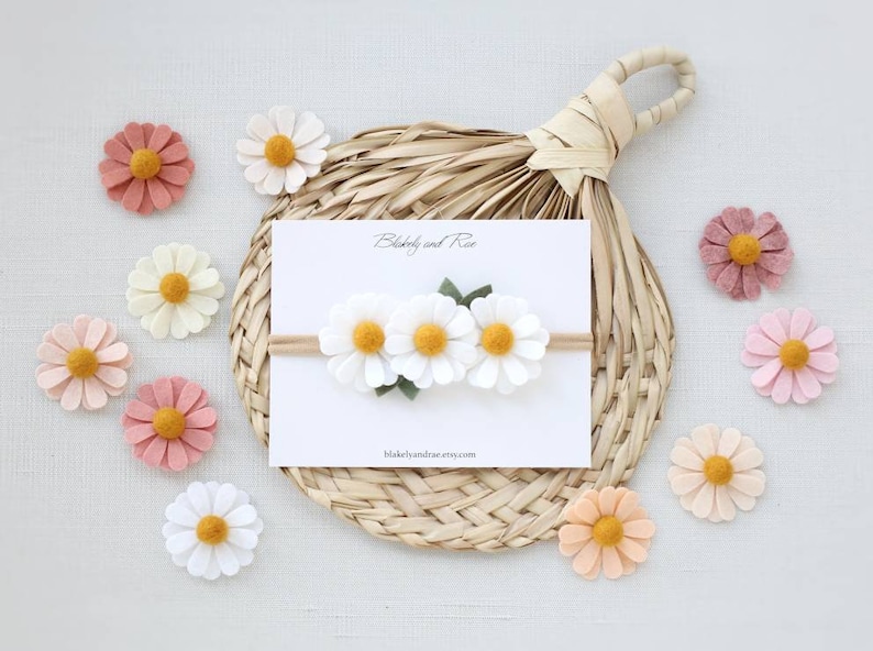 May include: A white felt headband with three white felt daisies and green felt leaves. The headband is on a white card with the text "Blakeley and Rae" and "blakeleyandrae.etsy.com". There are several felt daisies in various colors around the card.