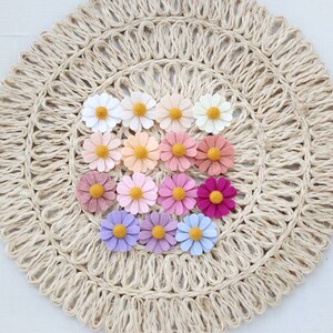 May include: A collection of felt flower embellishments in various shades of pink, purple, and white. The flowers are arranged in a circle on a natural woven mat.