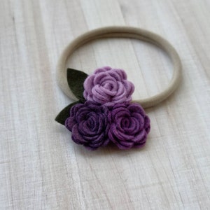May include: A purple felt flower headband with three flowers and green leaves. The headband is made of a nude elastic material.