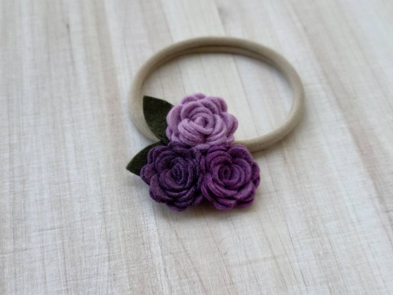 May include: A purple felt flower headband with three flowers and green leaves. The headband is made of a nude elastic material.
