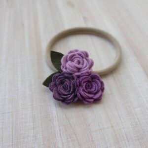 May include: A purple felt flower headband with three flowers and green leaves. The headband is made of a nude elastic material.