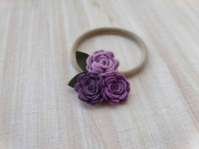 May include: A purple felt flower headband with three flowers and green leaves. The headband is made of a nude elastic material.