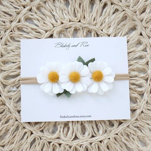 May include: A white felt headband with three white felt daisies with yellow centers and green felt leaves. The headband is on a white card with the text "Blakely and Rae" and "blakelyandrae.etsy.com".