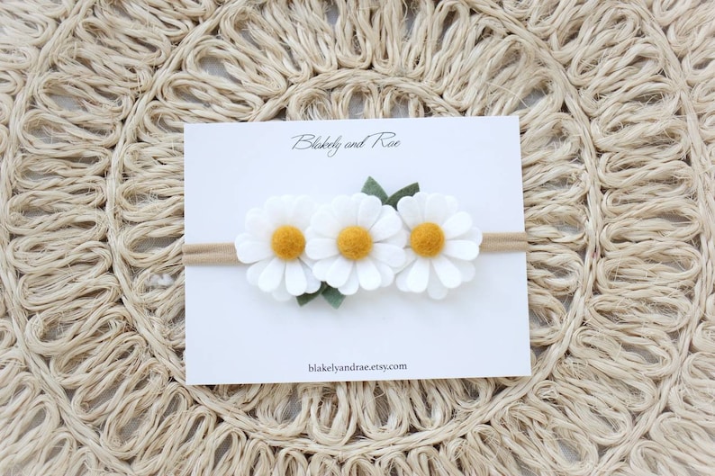 May include: A white felt headband with three white felt daisies with yellow centers and green felt leaves. The headband is on a white card with the text "Blakely and Rae" and "blakelyandrae.etsy.com".