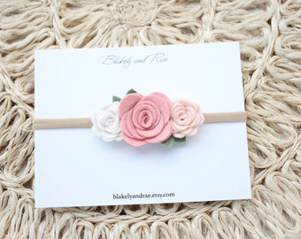 May include: A nude elastic headband with three felt flowers. The flowers are pink, white, and peach.