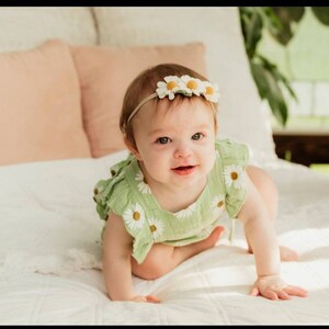 May include: A baby wearing a green and white daisy print onesie with a white daisy headband. The baby is crawling on a white bed.