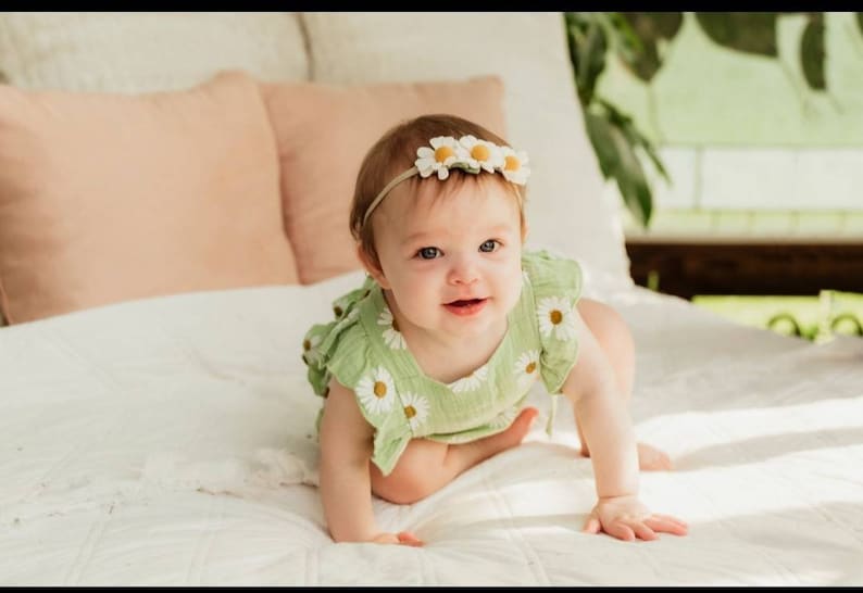 May include: A baby wearing a green and white daisy print onesie with a white daisy headband. The baby is crawling on a white bed.