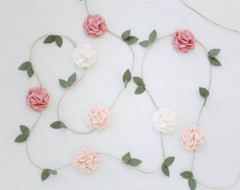 May include: A decorative garland featuring felt flowers in shades of dusty rose, pale pink, and white, interspersed with small green felt leaves attached to a thin, natural-colored twine string that winds across the frame in a loose, flowing pattern.