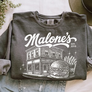 K&ouml;nnte beinhalten: Dunkelgraues Sweatshirt mit wei&szlig;er Grafik eines Restaurants, Burgers und Pommes Frites. Der Text "Malone's" befindet sich &uuml;ber dem Geb&auml;ude, mit "EST. 2015" rechts. Das Sweatshirt liegt gefaltet auf einer hellen Oberfl&auml;che.