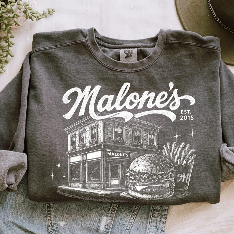 K&ouml;nnte beinhalten: Dunkelgraues Sweatshirt mit wei&szlig;er Grafik eines Restaurants, Burgers und Pommes Frites. Der Text "Malone's" befindet sich &uuml;ber dem Geb&auml;ude, mit "EST. 2015" rechts. Das Sweatshirt liegt gefaltet auf einer hellen Oberfl&auml;che.