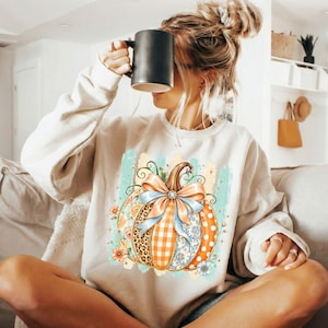 May include: A beige long-sleeve sweatshirt featuring a central graphic of a pumpkin decorated with various patterns including leopard print, orange gingham, and blue floral designs, topped with a large light blue bow and surrounded by small flowers.