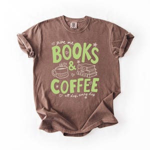 May include: A brown short-sleeve t-shirt featuring light green text that reads "give me BOOKS & COFFEE" along with white line art of a coffee mug, a stack of three books, small hearts, and flowers, plus the text "all day, every day".