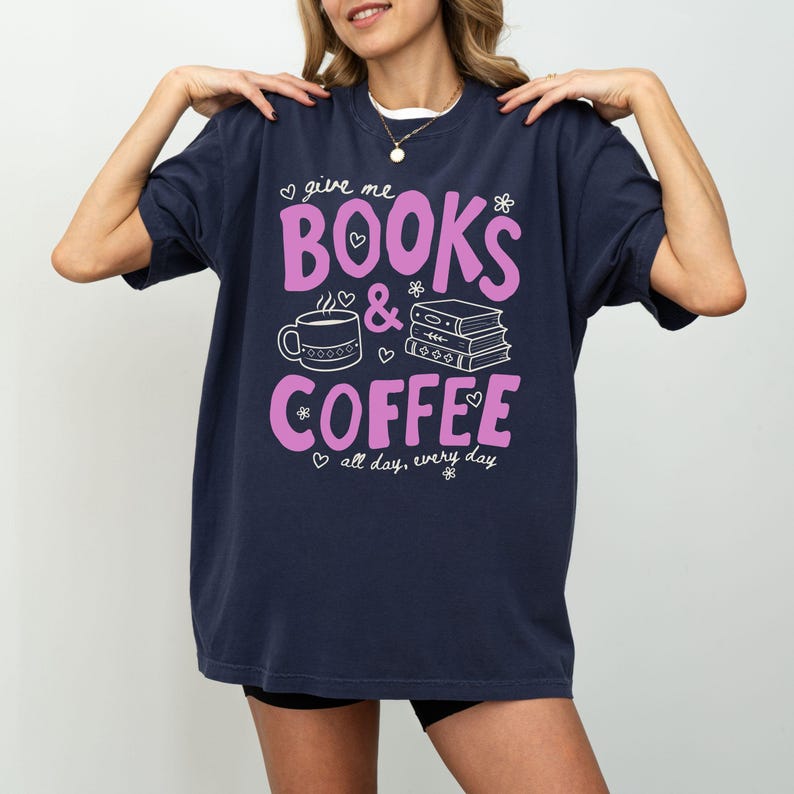 May include: A navy blue short-sleeve t-shirt featuring the pink text "give me BOOKS & COFFEE" with white line art of a coffee mug and a stack of books, accompanied by the white text "all day, every day" and small heart and flower icons.