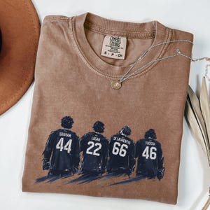 May include: A brown Comfort Colors t-shirt featuring a graphic of four hockey players with their jersey numbers: 44, 22, 66, and 46. The shirt has a crew neck and short sleeves. A silver necklace with a pendant is draped across the shirt.