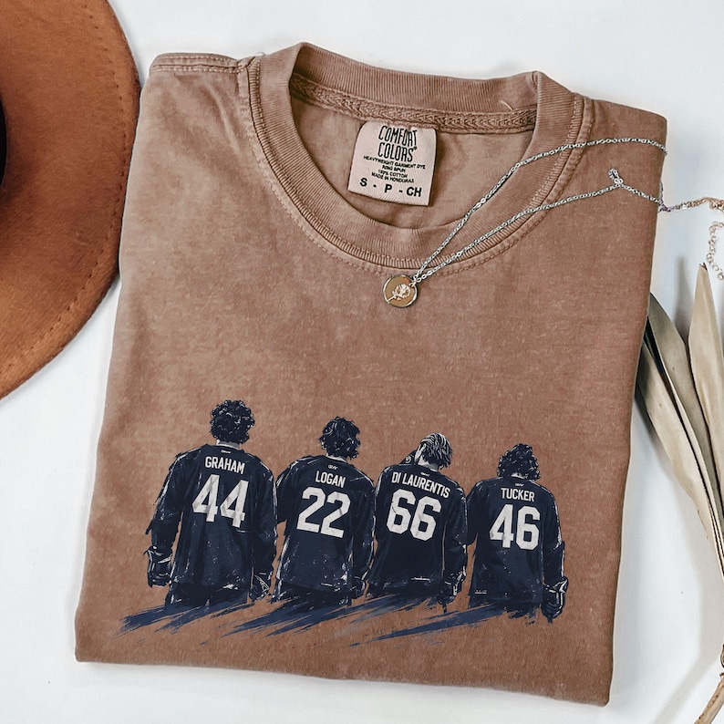 May include: A brown Comfort Colors t-shirt featuring a graphic of four hockey players with their jersey numbers: 44, 22, 66, and 46. The shirt has a crew neck and short sleeves. A silver necklace with a pendant is draped across the shirt.
