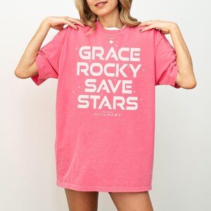 May include: A pink t-shirt with the words "GRACE ROCKY SAVE STARS" in white block letters. The text is centered on the front of the shirt, with the words stacked vertically. The brand name "PROJECT HAILMARY" is printed below the text.