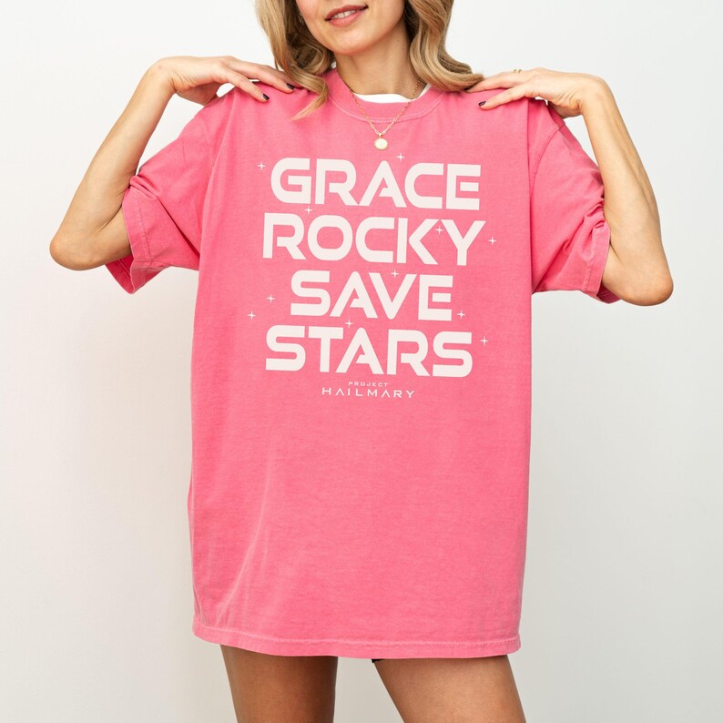 May include: A pink t-shirt with the words "GRACE ROCKY SAVE STARS" in white block letters. The text is centered on the front of the shirt, with the words stacked vertically. The brand name "PROJECT HAILMARY" is printed below the text.