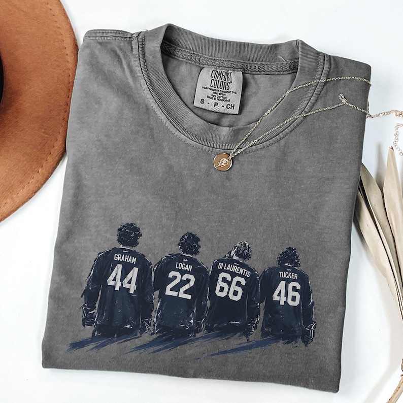 May include: Gray Comfort Colors t-shirt featuring a graphic of four hockey players with the names Graham, Logan, Di Laurentis, and Tucker, and the numbers 44, 22, 66, and 46. A silver necklace with a pendant is on the shirt.